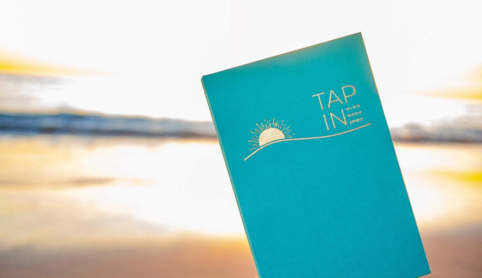 TAP IN wellness journal has a seafoam linen cover and a gold foil symbol of a sun and wave.